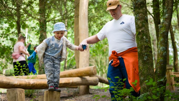 Boy balancing on wooden beam in the woods with the help of his dad