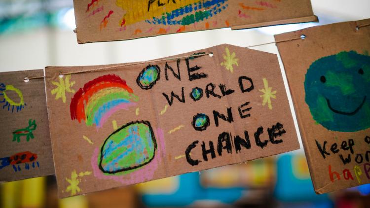 A cardboard placard hanging on a piece of string with 'one world one change' painted in black with another small paining of a rainbow and globe