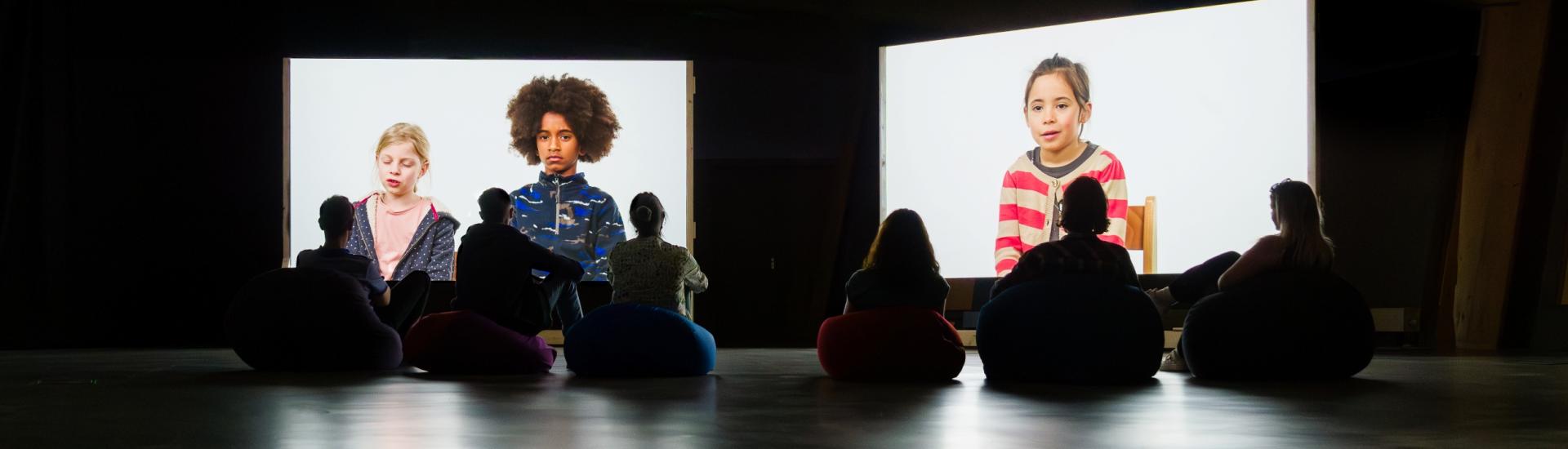 People in a dark exhibition space silhouetted against two screens showing a projection of children talking